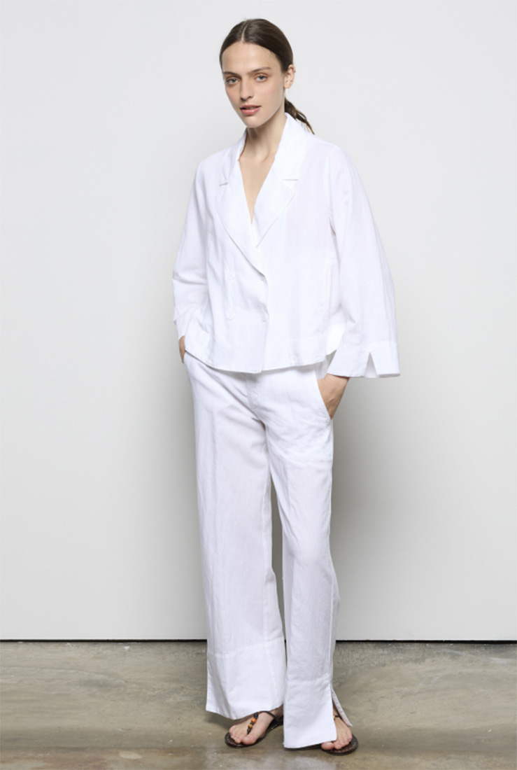 EU Woman in a white jacket and white pants stands with her hands in her pockets.