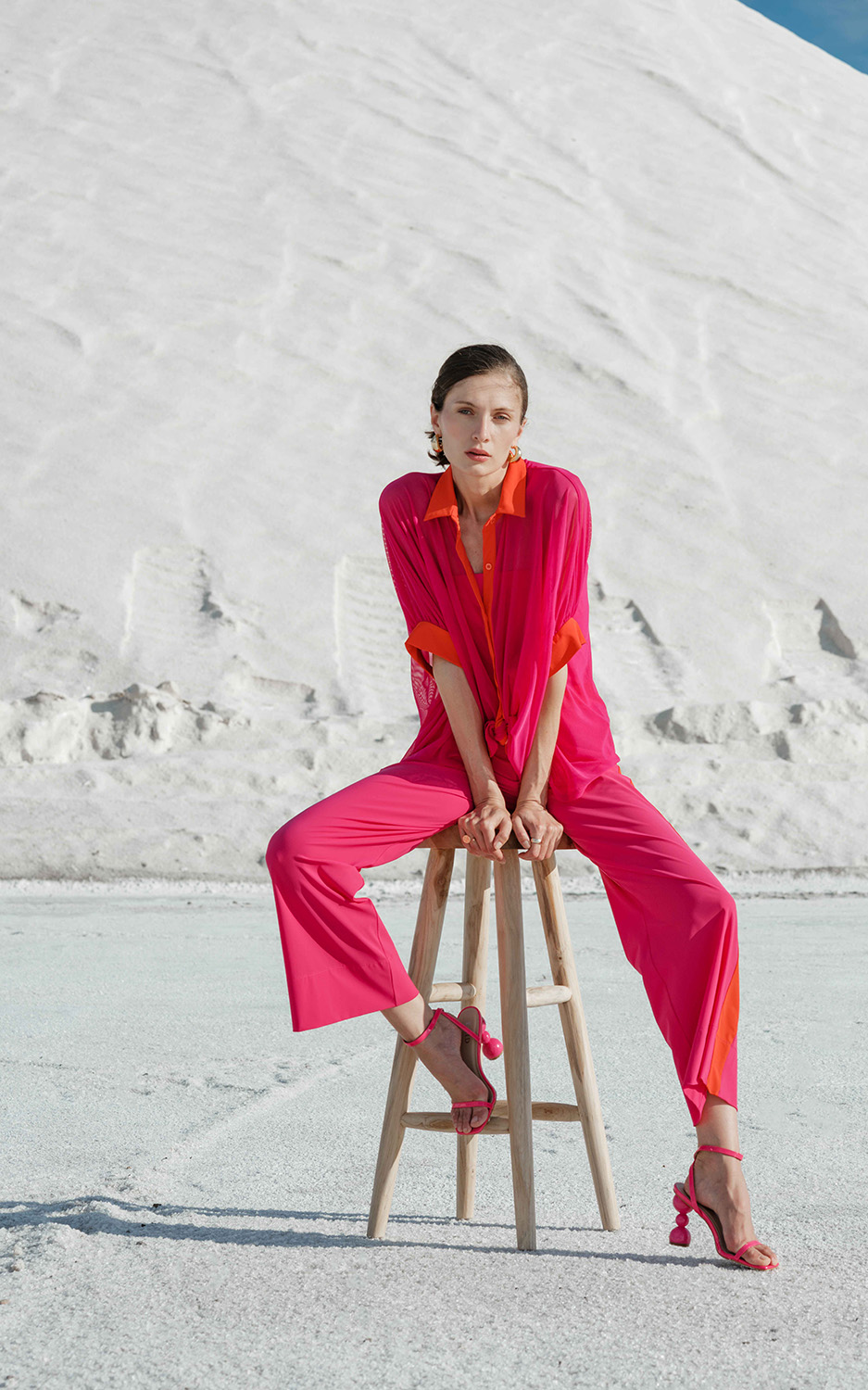 INDIES - SUMMER 25 - HD-72 Woman in bright pink loose fitting blouse and tailored trousers, seated on a stool.