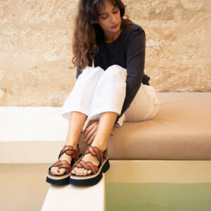 Woman in cropped jeans and high heel Homers sandals hugs her knees on a bench.